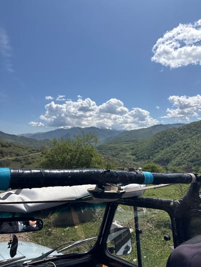 The image, likely taken from within a vehicle, shows a view of mountains and clouds against a blue sky. The location is inferred as Haghartsin, Armenia based on the presence of mountainous terrain and likely vegetation. The vehicle's frame, including a padded bar wrapped with black tape and blue accents, is visible. A white strap is connected to the bar. A side mirror and windshield wipers can be seen inside the car and a reflection of a person in the side mirror. The external landscape includes green slopes and cumulus clouds.