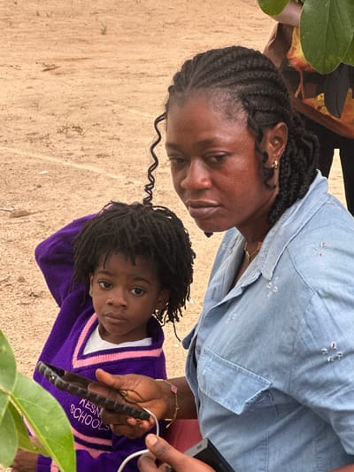 In Ganyingbo, Nigeria, a woman with braided hair and a light blue denim shirt sits next to a young child wearing a purple sweater. The woman is holding a dark-colored smartphone in her right hand, and a white charging cable is plugged into it. The child is looking directly at the camera. The background consists of a sandy, dirt-like ground with sparse vegetation and a blurred figure of another person partially visible behind the woman. The lighting suggests an outdoor setting, possibly during the daytime.