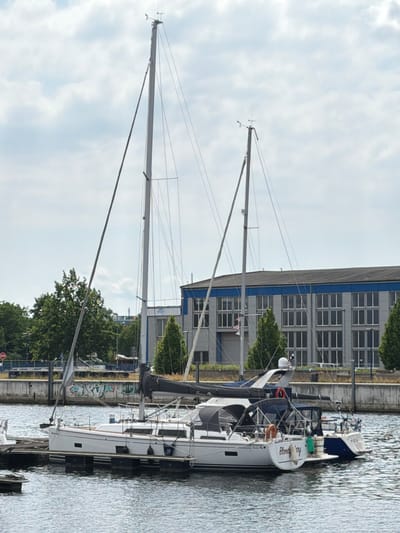 The image shows a marina in Wismar, Germany. Two sailboats are docked side-by-side. The larger sailboat on the left is white with two masts and a dark blue cover over part of the deck. The name "Bloody Mary" is visible on the stern of this boat. The smaller sailboat on the right is dark blue. Both boats are moored to a wooden dock. In the background, a large, modern building with a grey brick facade and a blue stripe runs along the top. Trees and a cloudy sky are also visible. The water appears calm. There are no people visible in the image.