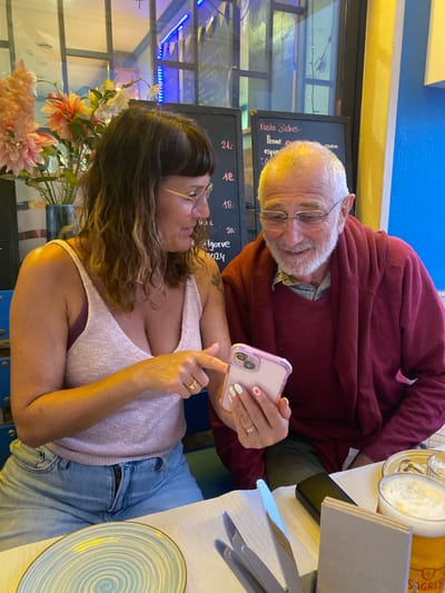In a restaurant setting in Olhão, Portugal, a woman with shoulder-length brown hair, glasses, and a light pink sleeveless top is showing a pink cell phone to an older man. The man, with white hair and a beard, wears glasses and a dark red sweater. He is smiling as he looks at the phone. The woman is wearing denim jeans, and the man is wearing dark trousers. They are seated at a table with a white tablecloth, cutlery, a blue and white swirled plate, and a glass of beer. In the background, a blackboard menu displays text that appears to be in Portuguese, and a vase of flowers is visible. The environment suggests an indoor dining area, possibly with outdoor elements visible through windows.