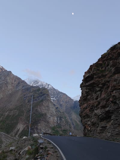 The image depicts a scenic mountain road in Gemur, India, at dusk. The sky is a pale blue with a sliver of the moon visible. To the right, a sheer rock face dominates the foreground, its rough texture and reddish-brown hues contrasting with the muted tones of the landscape. A winding asphalt road curves through the scene, marked by a white line. On the left, imposing mountains rise, their slopes covered in a mix of rock and sparse vegetation. Snow-capped peaks are visible in the distance, partially obscured by clouds. A solitary streetlamp stands on the side of the road, casting a faint glow. The overall impression is one of a remote and rugged mountain pass, bathed in the soft light of twilight.