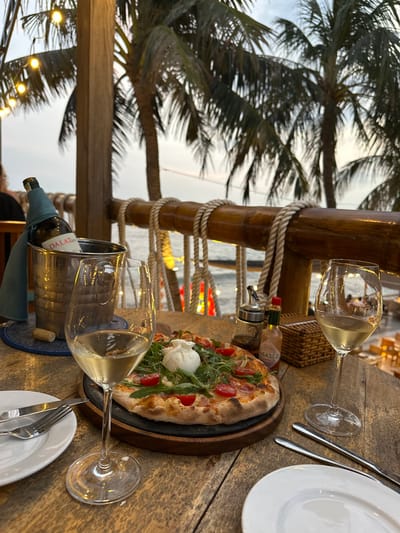 A dining table is viewed from a slightly elevated perspective in an outdoor restaurant setting in Thành phố Phan Thiết, Vietnam, at dusk. In the immediate foreground, on a rustic wooden plank table, a pizza with burrata, arugula, and cherry tomatoes rests on a dark round plate atop a wooden serving board. To the left of the pizza, two white wine glasses are positioned, one full and closer to the viewer, the other partially visible on the right. A chrome ice bucket containing a green-wrapped "DALAT" branded wine bottle sits on a blue and white coaster to the left of the wine glasses. Cutlery and a white plate are placed to the far left. To the right of the pizza, a small bottle of Tabasco sauce and a clear glass condiment dispenser with ground spices are present next to a small woven basket.
The midground features a prominent wooden railing adorned with thick white rope, creating a nautical theme. Beyond this railing, the dark expanse of the sea or ocean is visible. The background reveals multiple tall palm trees silhouetted against a pale sky, indicating twilight. String lights with visible bulbs are strung above, illuminating the dining area. Faint, warm lights are visible in the distant lower background, suggesting another section of the restaurant or a nearby area at a lower elevation. A single individual is partially visible sitting at a table in the far left background.