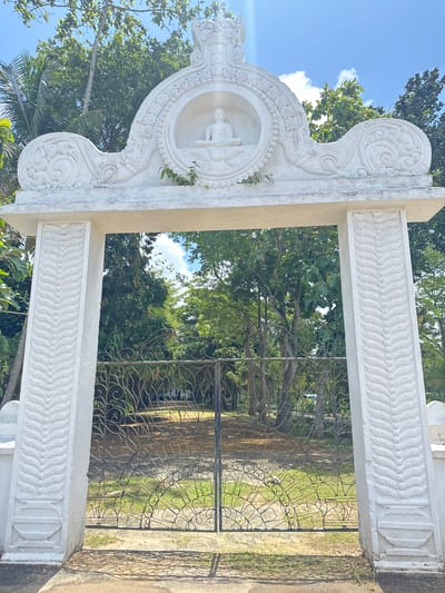 A low-angle, full-outdoor shot depicts an ornately decorated, white stone gate and fence in Walgama, Sri Lanka. The gate frame has intricate carvings, including a circular niche containing a seated Buddha statue at the top. The frame is supported by two vertical pillars also decorated with carved designs. A simple metal fence is behind the gate. Lush green trees and foliage fill the background, partially obscuring a glimpse of the sky.