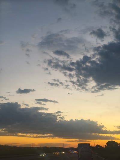 The image captures a sunset over a highway near Riba-roja de Túria, Spain. A dark cloud formation stretches across the sky, juxtaposed with the warm hues of the setting sun near the horizon. A small airplane is visible in the upper portion of the frame, traversing the sky. Below, the highway is lined with trees and features several vehicles, including a large truck in the foreground, its headlights illuminating the road. Power lines and transmission towers are also present in the background, characteristic of the Spanish landscape. The scene suggests a typical evening commute on a highway as daylight fades.