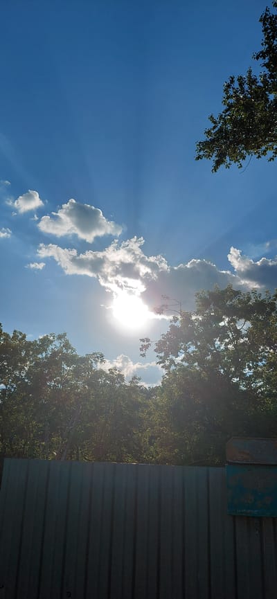 The image depicts a bright, sunny day with a clear blue sky and scattered white clouds. The sun is partially obscured by clouds, creating visible sunbeams. Lush green trees line the horizon, and a corrugated metal fence runs along the bottom of the frame. A rusty, blue mailbox is attached to the fence on the right side. The scene appears to be an outdoor setting, possibly a residential area or a park. The location is identified as Tashkazgan, Russia. There are no people or specific actions visible in the image.