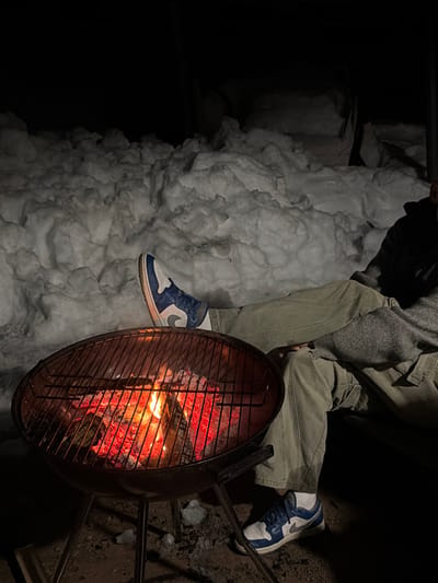 A nighttime scene in Ветеран Снт, Russia, shows an individual seated outdoors in a snowy environment. A round, black charcoal grill with glowing red embers and visible flames is positioned in the foreground. The grill features a metal cooking grate above the fire. The individual is wearing olive-colored cargo pants, white socks, and blue and white sneakers. Their left leg is extended with the foot resting near the top edge of the grill. A grey, textured garment covers a portion of their upper body. The background consists of large, uneven piles of snow under a dark sky. The ground directly around the grill is dark, indicating dirt or thawed earth.