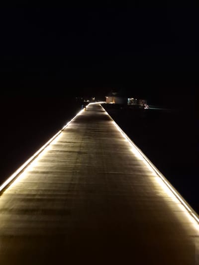 At night in Bodufolhudhoo, Maldives, a long, textured wall is illuminated with a linear light source along its top edge. The wall's surface appears to be composed of vertical elements. The surrounding area is dark. At a distance, a structure can be seen behind the wall.