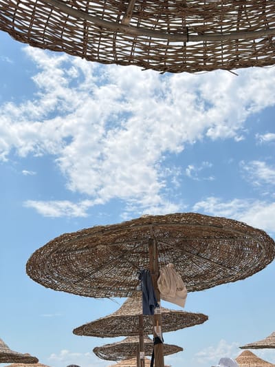 The image captures a low-angle view looking upwards towards a bright blue sky dotted with fluffy white clouds. The scene is dominated by several large, woven umbrellas, likely made of straw or reeds, which are characteristic of a beach setting. These umbrellas are angled to provide shade, and their woven texture is intricate and rustic.
On one of the umbrella structures, a few items of clothing are visible, draped over the supporting beams. One piece appears to be a light-colored fabric, possibly a towel or shirt, while another is a darker, folded garment. The presence of clothing suggests that people have been using these umbrellas for shade.
The overall impression is one of a sunny day at the beach. The lighting suggests it is daytime, likely late morning or early afternoon, given the brightness of the sky and the shadows cast by the umbrellas. The weather appears clear and pleasant, ideal for a beach outing. The perspective from below emphasizes the scale and structure of the umbrellas against the vastness of the sky.
While there are no explicit people visible in the image, the scene implies human presence and activity. The setting is clearly an outdoor recreational area, most likely a beach, given the type of umbrellas. The location context provided, Ofrynio Beach, Greece, reinforces this interpretation. The image evokes a relaxed, summery atmosphere. There is no visible text in the image.