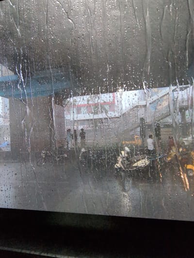This image is taken from inside a vehicle, looking out through a rain-streaked window at an urban street scene in Hyderabad, India. The window is covered in numerous raindrops, some large and some small, which distort the view of the outside. The ambient light suggests it is daytime, possibly overcast or raining heavily.

Beneath a blue overhead structure, possibly an overpass or flyover, a street is visible with several people and vehicles. The area appears to be a busy junction or intersection. There are at least four individuals visible, dressed in casual clothing. One person in the center is wearing a white shirt and dark trousers. Another person to their right is wearing a dark shirt and pants. Further to the right, a person in a red top is visible. To the left, some figures are also present.

Various vehicles are present on the street. A yellow auto-rickshaw is parked prominently in the middle of the scene. Several motorcycles or scooters are also visible, some parked and others potentially in motion. A yellow vehicle, possibly a small truck or construction equipment, is also present in the background.

The background shows buildings with signs, typical of a commercial area in India. One sign appears to have red lettering. A pedestrian walkway or elevated structure is visible behind the vehicles. The overall impression is a street scene in Hyderabad during rainfall, with the rain creating a visual filter.