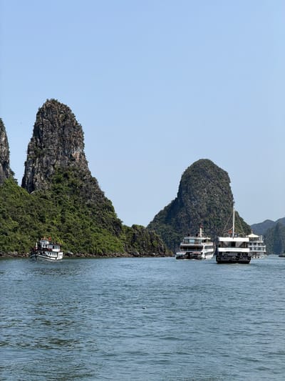 This image captures a serene bay scene in what appears to be Tuan Chau Ward, Vietnam, characterized by majestic limestone karsts rising from the water. The sky is a clear, light blue, suggesting a bright and sunny daytime. The water, a calm blue-green, shows gentle ripples across its surface.

The landscape is dominated by several large, steep-sided karsts, densely covered in rich green vegetation. These natural formations stretch across the midground and background, creating a dramatic and picturesque setting.

Several cruise boats are visible on the water, indicating active tourism or leisure. Two larger, multi-decked white boats with dark hulls are positioned closer to the karsts in the middle-right of the frame. One of these boats clearly displays the text "HERITAGE LUXURY CRUISE" on its side, identifying it as a tourist vessel. Another smaller white boat is visible further to the left, near another karst. The boats appear to be either cruising or anchored, suggesting a tranquil exploration of the bay. No specific people are discernible, but their presence is implied by the active cruise boats. The overall ambiance is one of natural beauty and peaceful exploration under clear weather conditions.