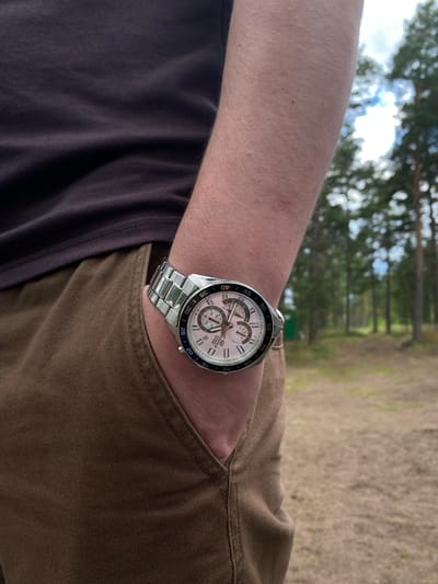 A person's arm wearing a silver watch with a white face and multiple subdials is positioned with their hand in the pocket of brown pants. The person is wearing a dark t-shirt. The background is a blurred forest with tall trees and a bright sky. The scene appears to be outdoors in a natural setting, possibly a park or wooded area. The location is Baltiiskoe, Russia.