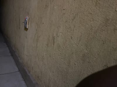 The image shows a close-up of a rough, textured concrete wall in dim lighting. To the right of the wall, there are gray, rectangular ceiling tiles. A small, rectangular electrical outlet is visible, secured to the wall. There are no people visible. There is no indication of the location being Sabo, Nigeria.