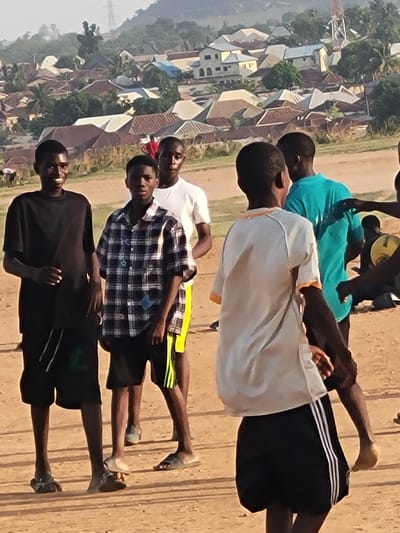 A group of four young men stand outdoors in Ibo, Nigeria. They are facing forward, arranged in a line, with the background displaying buildings and a rural cityscape.
The first individual is wearing a black shirt and dark shorts with a white stripe, and sandals on his feet. The next person is clad in a blue and white plaid shirt with yellow striped shorts and dark sandals. The third figure has a white shirt and black shorts. The fourth individual wears a light blue shirt with black shorts bearing white stripes. The first three are barefoot. All four subjects appear to be young men. The setting suggests a rural or semi-rural region within the city.