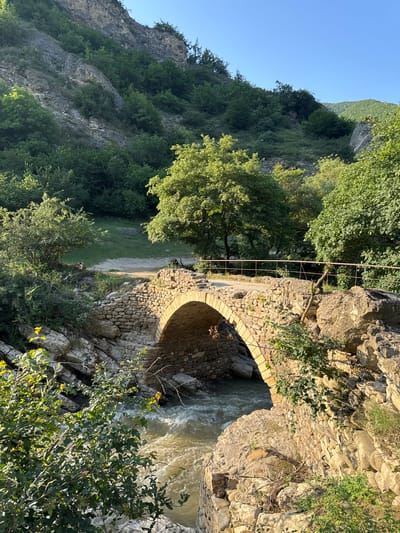 The image depicts a stone arched bridge crossing a flowing river in a mountainous, forested region. The bridge appears old and constructed from rough-hewn stones. A metal railing runs along the top of the bridge. The surrounding environment is lush with green trees and vegetation, with rocky cliffs visible in the background. The river is wide and appears to be moving quickly, with some white water visible. The sky is clear and blue. The scene appears to be set in the daytime, with sunlight illuminating parts of the bridge and river. The location is identified as Khanag, Russia. There are no people visible in the image.
