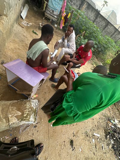 The image depicts an outdoor scene in Iriebe, Nigeria, showing five individuals gathered on a dirt-and-sand ground amidst various objects and structures.

In the foreground left, a male with dark skin and curly hair, shirtless with a light green towel draped over his left shoulder, wears red shorts and dark slippers. He sits on a low, purple wooden structure and holds a rolled white paper in both hands, examining it. To his right and slightly behind, a male with lighter skin, dark hair, and a beard wears a light-colored hooded tracksuit and white slippers. He sits on a dark object, resting his hands near his face and observing the first male. Adjacent to him, a male with dark skin and a receding hairline wears a red short-sleeved t-shirt with a Manchester United logo and dark shorts. He sits on a dark object, with his left arm resting on his bent left knee, also observing the first male. In the foreground right, a person with dark skin, wearing a bright green long-sleeved garment and dark slippers, holds a white smartphone with both hands, focusing on its screen. A black shoulder bag rests beside this individual. Feet in dark sandals, possibly belonging to the photographer or another unseen person, are visible at the bottom edge of the frame.

The environment includes a worn concrete block wall on the left. In the background left, a makeshift wooden structure draped with a blue tarp and hanging clothes (red, pink, yellow) is visible. A green glass bottle lies on the ground near the first male. The ground itself is uneven, sandy, and scattered with small pieces of debris. Behind the group, green vegetation and a taller concrete block wall topped with barbed wire are present, suggesting a boundary or enclosure. The sky is overcast.