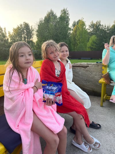 Three children are sitting on a yellow bench, wrapped in bathrobes: a girl in pink in the foreground, a girl in red with a package of Zerfer Duo marshmallows in the middle, and a girl in white in the background. An adult is visible on another bench in the upper left of the frame. The location is outdoor with trees and building visible in the background, Новая Чернушка, Russia.