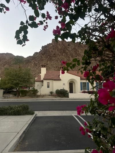 This image displays a residential street in La Quinta, United States, showcasing a property identified by the address 77752. The central subject is a cream-colored stucco house featuring a red terracotta tile roof, multiple chimneys, an arched wooden main entrance, and windows with blue louvered shutters. An outdoor air conditioning unit is visible near the house's front. The residence is positioned opposite a dark asphalt street, which includes a concrete sidewalk and a paved parking area marked with two faint white lines. Landscaping in front of the house and along the street consists of various green shrubs and low vegetation. To the left of the house, a tall palm tree and a smaller deciduous tree are present. A dark bench is partially visible on the far left. The background is dominated by rugged, brown, rocky mountains beneath a light gray sky. The foreground is partially obscured by vibrant magenta-pink bougainvillea flowers and dark green leaves. No individuals or active interactions are depicted.