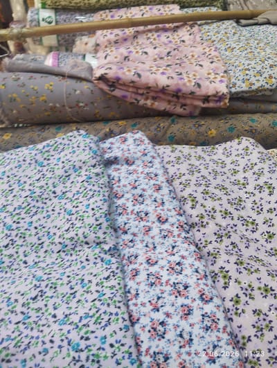 The image shows a close-up of folded fabric rolls with floral patterns, displayed in what appears to be a market or shop in Istaravshan, Tajikistan. The fabric patterns include small flowers in various colors such as pink, purple, blue, green, and yellow, against light-colored backgrounds like pink, gray, and white. The rolls are stacked, with the closest ones featuring intricate floral designs. A wooden rod is visible horizontally across the upper portion of the image, suggesting a display rack. The lighting appears to be artificial, possibly from overhead lights in a covered market. The bottom right corner of the image contains a timestamp "22.06.2026 11:23". There are no people or any specific environmental features of Istaravshan city visible in this close-up shot of the merchandise.