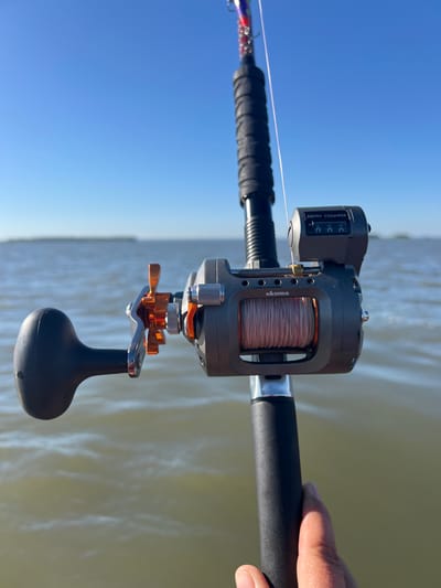 An over-the-shoulder, medium shot captures a fishing rod with a reel, held by a person's hand, angled upwards against a clear blue sky. The reel, a gray and orange Okuma model with a depth counter, is in focus, its line visible. The background shows a calm body of water with a faint landmass on the horizon. The image appears to be taken outdoors, possibly on a boat. The location in Novopichugovo, Russia is not discernible from the visual elements of the image.