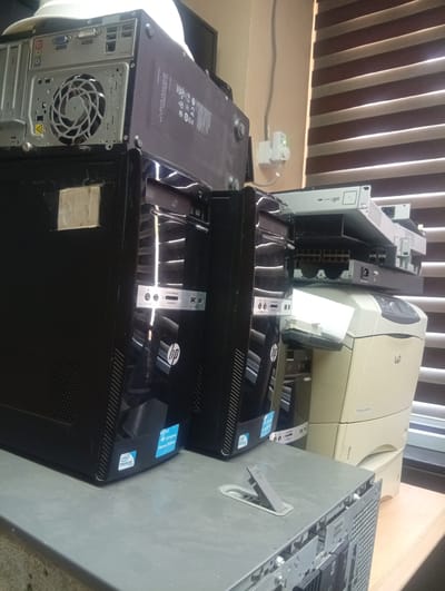 The image shows a cluttered desk area with various electronic devices. In the foreground, a grey metal casing of a computer, possibly a server, is partially disassembled, revealing its internal components and ports. To the left, two black HP Compaq desktop computers are stacked. The front panel of the bottom computer displays an optical drive, USB ports, and audio jacks, with an "HP" logo prominently visible. A blue sticker indicating "HP Compaq Home Edition" is affixed to its side. Another sticker on the bottom left shows an "Intel Pentium" logo.
Behind the desktop computers, a beige HP Laserjet printer is visible. Stacked above the desktop computers are several network devices, including a white Ubiquiti UniFi switch and other rack-mounted equipment. A white power adapter with a green LED light is plugged into a wall outlet on the left side of the image. The background features beige-colored walls and horizontal blinds on a window. The overall scene appears to be an office or IT environment. The location is identified as Shiroro, Nigeria, suggesting this is a snapshot of technology infrastructure within that area.