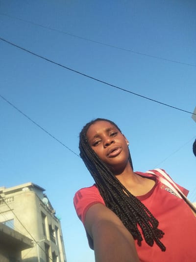 A young dark-skinned female is depicted in Cotonou, Benin, captured in a low-angle selfie. She occupies the lower-center and right of the frame, looking directly at the camera with a neutral expression and slightly parted lips. Her hair is styled in long, black box braids. She wears a plain red short-sleeved t-shirt, featuring a faint dark emblem or stripes on the right shoulder, and a white and red striped bag strap crosses her right shoulder. A small, clear stud earring is visible in her right ear. The background primarily consists of a clear, bright blue sky, with multiple thin, dark utility lines traversing it diagonally. In the lower-left quadrant, a multi-story concrete building is partially visible, featuring windows, balconies, and decorative balustrades on its upper floors. Two satellite dishes, one white and one light blue, are mounted on the upper exterior of this building. The scene is illuminated by natural daylight.