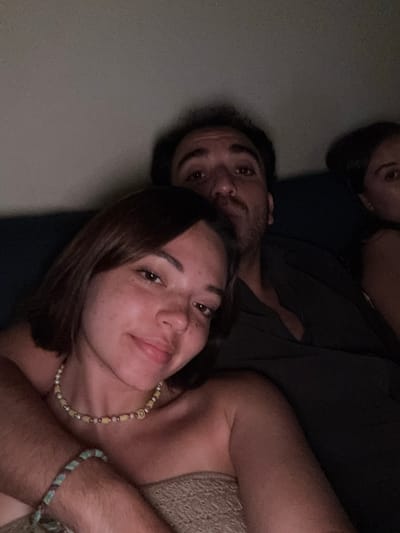 The image shows a close-up, selfie-style photograph of three individuals. In the foreground, a young woman with shoulder-length brown hair and a necklace with a smiley face charm smiles at the camera. She is embracing someone with her left arm. Behind her, a man with dark hair and a mustache looks towards the camera. To the right of the man, the face of another woman is partially visible. The individuals appear to be indoors, possibly in a dimly lit room. The image does not contain any information about the location in Chevy Chase, United States of America.