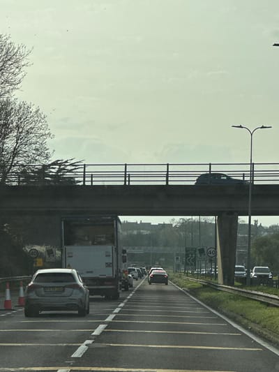 The image displays a multi-lane road experiencing traffic congestion under a concrete overpass in Basildon, United Kingdom. This location is identified as the A127 (Southend Arterial Road) due to visible directional signs for "M25" and "A127" on an overhead gantry further down the road, coupled with a "40" mph speed limit.
The perspective is from within a vehicle, facing eastbound. In the foreground, two orange traffic cones are positioned on the left shoulder. A silver Mercedes-Benz A-Class hatchback (UK registration PX70 JXG) is stationary or slow-moving in the left lane. In the adjacent lane, a large white box truck with rear red and white chevron markings is situated directly beneath the overpass, also stationary or slow-moving. A queue of additional vehicles, including a dark sedan with illuminated taillights, extends into the distance, indicating traffic.
The concrete overpass spans the road, featuring a black metal railing on its upper edge. A dark SUV or similar vehicle is observed moving across the overpass from left to right. A tall black streetlight with a double-headed luminaire is positioned on the right side of the overpass. On the left side of the road, bare deciduous trees and evergreen shrubs are visible. Road signs include a circular "40" mph speed limit sign and, further ahead, an overhead gantry displaying directional signs for "M25," "A127," and a congestion warning. The road surface features distinctive yellow horizontal rumble strips and white lane markings. The right side of the carriageway has a grassy verge and a metal crash barrier. The sky is overcast and pale grey. Distant urban structures are faintly visible on the horizon.