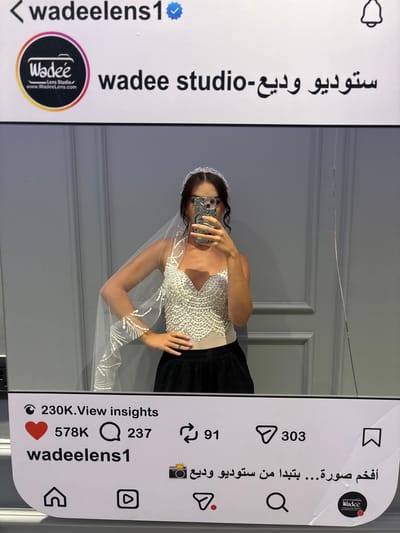 An Instagram post simulation is displayed, featuring a profile picture, username, and engagement metrics. The profile picture is a circular logo with the text "Wadee Lens Studio" and "www.Wadeelens.com". The username "wadeelens1" is visible, followed by Arabic text that translates to "Wadee studio". Below this, engagement icons show 230K views, 578K likes, 237 comments, and 303 shares. A caption in Arabic, translating to "The most amazing photo... starting from Wadee Studio", is also present, along with icons for home, play button, search, and a bookmark. The simulated post is overlaid on a photograph of a woman in a wedding veil and a sparkling top, taking a selfie in a mirror. The background is a wall with vertical paneling. This image was taken in Sweileh, Jordan.