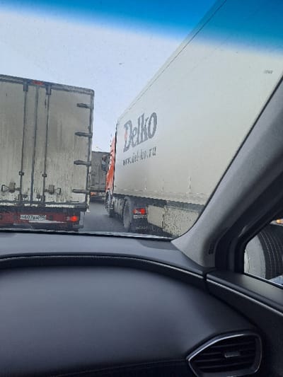 The image depicts a traffic queue of large commercial trucks on a multi-lane road in Obukhovo, Russia, observed from inside a stationary or slow-moving vehicle.
In the foreground, the dark dashboard, lower windshield, and right-side A-pillar of the observer's vehicle are visible. The dashboard features a textured surface, and a silver-accented air vent is partially seen on the far right. The top portion of the windshield shows a blue tint.
Ahead and to the left, the rear of a dirty, light-colored box truck is visible. Its two red taillights are illuminated. A Russian license plate, "H607BA|750", is affixed to its lower rear. The "750" region code corresponds to Moscow Oblast.
To the right, immediately adjacent to the observer's vehicle, is the right side and rear of a white box truck featuring "Delko" and "www.del-ko.ru" in dark blue lettering on its side. The orange cab of this truck is partially visible. Its tires and lower body are heavily soiled with dirt and grime. A partially visible Russian license plate, "H220B", is on its rear.
Further down the road, additional trucks are queued in both visible lanes. The road surface appears wet or damp. The sky is overcast, indicating a dull daytime environment. No individuals are visible within the frame.