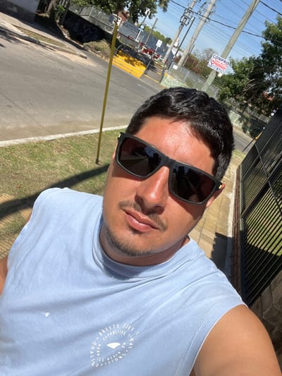 A male individual, facing the camera for a selfie, is positioned outdoors on a clear, sunny day. He has dark hair, a mustache, and light stubble on his chin. He wears dark rectangular sunglasses with subtle yellow accents on the frames and a light blue sleeveless t-shirt featuring a white circular graphic. The graphic contains text including "BREEZE SURF CO. ESTABLISHED SYDNEY WE PROVIDE QUALITY SURF & BEACH PROVISIONS BORN." His skin appears tanned.
The background shows a suburban street environment within Partido de Quilmes, Argentina. To the subject's immediate right is a dark metal fence, bordering a light-colored sidewalk. To his left is a strip of green grass, followed by an asphalt road. Across the road, a residential area with houses and trees is visible. Prominently in the midground, a railway crossing is present, characterized by raised yellow and white striped barriers. Multiple utility poles with overhead wires stand in the background. Attached to one of these poles is a white and red sign reading "COMPRO SU AUTO" ("I buy your car") followed by phone numbers, partially obscured, beginning with "11".