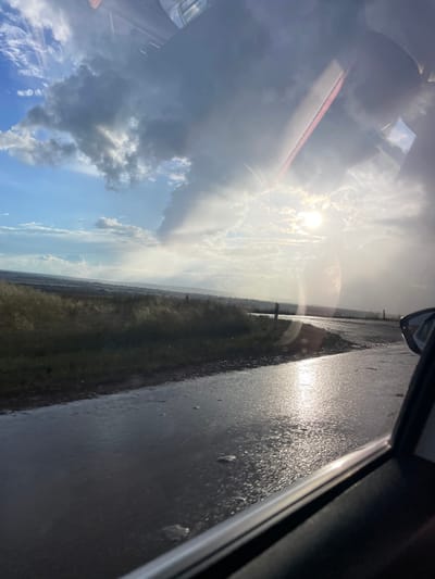 The image depicts a scenic view from inside a vehicle, likely in Kipchak-Askarovo, Russia, on a partly cloudy day. The sun is partially obscured by clouds, casting a bright glare across the scene and reflecting off a wet road. The road appears to be asphalt and is covered in puddles and streaks of water, suggesting recent rain. On the left side of the road, there is a grassy or reedy field stretching towards rolling hills in the distance, which are hazy and have a muted, earthy tone. The sky is a mixture of bright blue and dramatic, dark gray clouds. The sun's rays are visible breaking through the clouds, creating a dramatic lighting effect. The image is taken from a low angle, emphasizing the sky and the wet road. A portion of the car's interior, including a window frame and part of the dashboard, is visible in the foreground, framing the view of the landscape. There are no people visible in the image, and the scene appears to be in a rural or semi-rural setting.