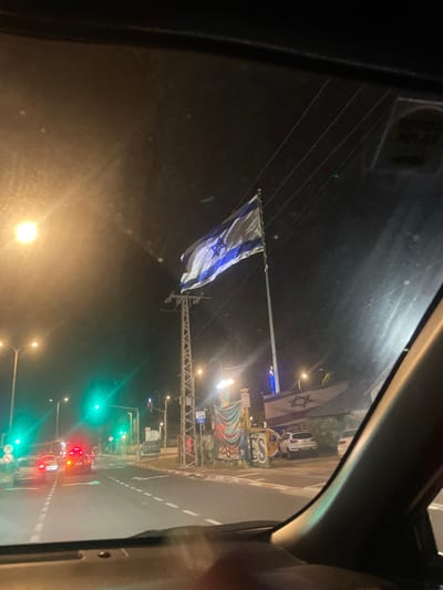 A nighttime view from inside a vehicle. The Israeli flag hangs on a pole over a street with traffic lights showing green. Another Israeli flag is visible in the background. Streetlights illuminate the road, and a building with graffiti is visible on the right. The reflection of the interior of the car is also visible in the window.