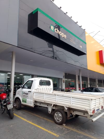 A white flatbed truck is parked on a street in front of a commercial establishment in Cuiabá, Brazil. The truck has "e chope" logo and phone number on its side door. A red motorcycle is parked to the left of the truck. The commercial establishment features a black facade with a green stripe and a large illuminated sign that reads "e chope". The establishment also has a yellow facade and a red rectangular accent on its right side. The sky is overcast.