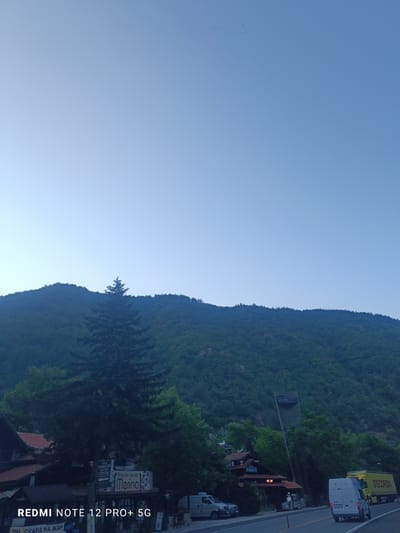 The image captures a daytime scene in the vicinity of Stara Kresna, Bulgaria, with a focus on the natural landscape and a roadside area. A dense, tree-covered mountain range dominates the background, stretching across the upper half of the frame. The sky above is a clear, pale blue, with a distant flock of birds visible in a V-formation. In the foreground, a paved road runs horizontally, with a white van and a yellow truck parked on it. To the left of the road, a building with a tiled roof and a sign that reads "Merina" is visible, alongside other structures that appear to be part of a village or small town. A large evergreen tree stands prominently in the middle ground, between the road and the mountain. The lighting suggests it is either early morning or late afternoon, with soft light illuminating the scene. The overall impression is one of a scenic, somewhat rural location in Bulgaria.
