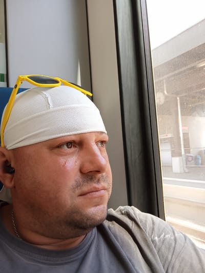 The media file is a close-up, waist-up photograph of a bald man wearing a white skullcap and yellow sunglasses perched on top of it. He is looking out of a window, his expression appearing contemplative. The man has a light beard and is wearing a grey t-shirt with a silver chain around his neck. A black earbud is visible in his left ear. The setting appears to be a mode of transport, possibly a train, with a window showing a blurry outdoor scene. The light suggests it is daytime. The background outside the window shows a platform or station with some structures that are out of focus. There is no visible text in the image.