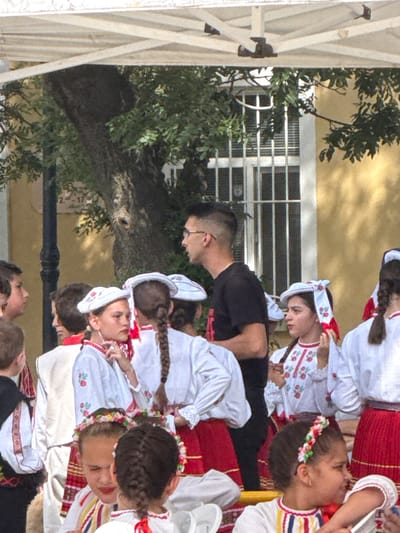 In Pordim, Bulgaria, an outdoor event is underway beneath a white canopy. A group of children, some wearing traditional Bulgarian attire, are gathered. One young girl in the foreground wears a white blouse with embroidered red floral patterns and a white cap adorned with similar embroidery and red ribbons. She has dark braided hair and is looking to her right. Behind her, other children are also dressed in traditional clothing, featuring white blouses with red embroidery and red skirts. A young boy in the background is wearing a black t-shirt. The scene takes place near a building with yellow walls and a window with white blinds. A large tree with dark bark and green leaves is visible behind the children. A black lamppost stands to the left of the frame.