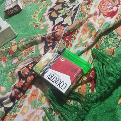 The image shows a close-up, high-angle view of a pack of "Country International" cigarettes and a green lighter resting on a green fabric with a colorful floral pattern. The fabric appears to be a scarf or shawl, evidenced by the fringed edges visible on the right side of the image. The "Country" cigarette pack is red and white, with text that includes "PERINGATAN" (Indonesian for warning), indicating health warnings typically found on tobacco products. The lighter is translucent green. The background fabric is densely patterned with various floral motifs in shades of green, orange, red, and yellow, suggesting a decorative textile. The overall lighting is dim. The image composition focuses on the items on the fabric, with no discernible context for their location within Kedung Dowo, Indonesia. There are no people or actions depicted.