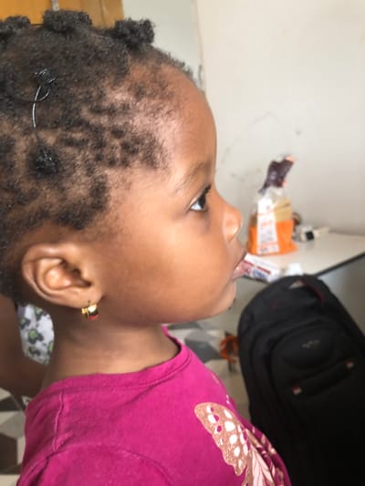 The image depicts a young, dark-skinned child, likely female, in a residential setting in Ilishan, Nigeria. The child is presented in a close-up profile view, facing towards the right with her head slightly elevated and eyes directed upwards. Her dark, short hair is styled in small knots, with a thin dark wire or thread visible in one section above her left temple. She wears a bright pink short-sleeved top adorned with a light-colored butterfly motif on the right chest area. A small, gold-colored hoop earring with a red accent is present in her left earlobe. To the far left, a portion of another individual's limb, dressed in a white garment with dark patterns, is partially visible behind the child. The background consists of a plain, light-colored wall. On a surface in the mid-ground right, a brown and orange package is visible, along with a darker, indistinct object behind it. A black backpack with red accents is located on the lower right side of the frame.