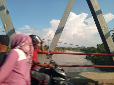 The image captures a view from a bridge over a river in Kulukuta, Indonesia. Two people are on a motorbike, with the driver wearing a helmet and a red shirt, and the passenger in front wearing a purple hijab and a pink garment. They are captured in motion, indicated by motion blur. The bridge's structure, with its metallic supports, is visible, along with red railings. The river flows through a landscape populated by lush green trees and vegetation, suggesting a tropical environment. The sky above is blue with scattered white clouds. Power lines are visible stretching across the frame.