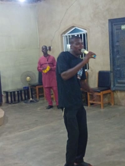 In a room, a man dressed in black shirt and pants stands holding a microphone and appears to be speaking or singing. Behind him to the left, another man in a red outfit with a tambourine stands in the background. An electric fan is visible in the distance near a bench. A window with bars and a dark door frame the scene. The room appears simple with light-colored walls.