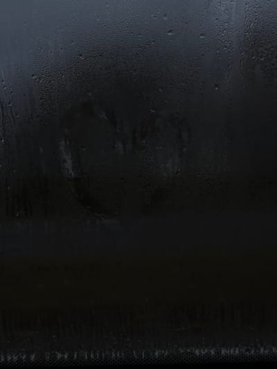 The image displays a close-up view of a glass surface, identified as a vehicle windshield or window, heavily covered with condensation and numerous small water droplets. A heart shape has been wiped into the condensation, centrally located in the upper middle section of the frame, revealing a slightly clearer outline against the misty surface. Various sizes of water droplets adhere to the glass, with some coalescing into vertical streaks and rivulets, particularly on the left. The background visible through the glass is uniformly dark and indistinct, showing no discernible features. A textured horizontal band, consistent with a vehicle's dashboard or defroster grid, is visible along the bottom edge of the frame. The scene is located in Пашкаўскі сельскі Савет, Belarus. No individuals are depicted, and the only observed interaction is the implied drawing of the heart. The environment suggests high humidity or a significant temperature differential causing the condensation.
