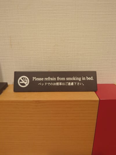 A dark rectangular sign with white text and a no-smoking symbol is placed on a light brown wooden surface. The sign reads "Please refrain from smoking in bed." and below it in Japanese characters "ベッドでのお煙草はご遠慮下さい。" indicating "Please refrain from smoking in bed." The background is a textured light beige wall. To the right of the wooden surface is a portion of a bright red object. The scene appears to be indoors, likely in a hotel room in Narita, Japan.