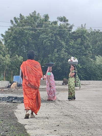 In Walsawangi, India, a woman in an orange sari walks away from the camera on a dirt and gravel path. Behind her, a tan dog sniffs the ground near a blue container. To her right, two women stand. The woman on the left wears a pink and white patterned sari and carries a dark bowl on her head. The woman on the right wears a green and black checkered sari and carries a bowl filled with various items on her head. She is smiling and touching her face. In the background, lush green trees form a dense canopy against a hazy sky. The environment appears to be an outdoor, possibly construction or work site.
