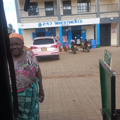 The image shows a view from inside a vehicle of a woman standing near a street in Sheywe ward, Kenya. The woman wears a colorful floral top, a green and blue patterned skirt, and an orange headscarf. Behind her, a pink Audi SUV with license plate "KDQ 481W" is parked in front of a building with the sign "247 INVESTMENTS, WE VALUE YOU KAKAMEGA BRANCH". Various items, including mattresses, gas cylinders, and plastic basins, are placed in front of the building. Two motorcycles with license plates "KNFU 4778" and "KMBK 4022" are parked to the right of the car. A "TO LET" sign and a "M PESA" advertisement are also visible near the building. The street is paved with interlocking concrete blocks. Parts of the vehicle frame from which the photo is taken are visible on the left and right sides of the image.