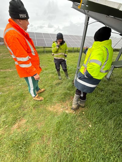 An outdoor scene at a solar farm in Southampton, United Kingdom, is depicted under an overcast sky. Three male individuals are positioned in a grassy field with patches of exposed earth. In the foreground left, a man wears an orange high-visibility jacket, green sweatpants with a white stripe, brown work boots, and a black beanie bearing "ROBINSON & LAWLOR" text; he holds a lit cigarette in his right hand. In the mid-ground center, another man, wearing a yellow high-visibility jacket, dark work trousers, green wellington boots, and a black beanie with ear flaps, holds a mobile phone to his right ear, speaking. In the foreground right, a third man, wearing a yellow and dark blue high-visibility jacket, dark work boots, and a black beanie, is partially obscured by the metallic frame of a solar panel. He stands with his back to the viewer, gesturing with his right hand towards the underside of the solar panel structure. Rows of ground-mounted solar panels extend across the background.