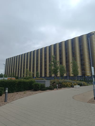 A wide, low-angle exterior shot captures a modern, multi-story car park with a distinctive yellow and black patterned facade. The building dominates the frame, stretching horizontally across the image. The facade is comprised of numerous overlapping horizontal slats, creating a wave-like pattern. Green trees and shrubs are visible at the base of the building, adding a touch of nature to the urban landscape. In the foreground, a paved walkway curves to the right, bordered by gravel and low-level lighting bollards. A white ambulance vehicle is partially visible to the left, behind a hedge. The sky is overcast and grey. The location is the Cambridge Biomedical Campus in Cambridge, United Kingdom.
