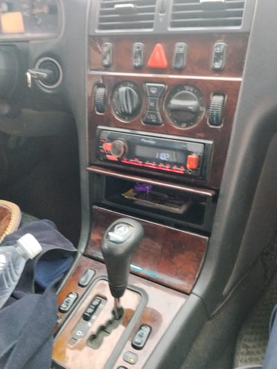 The image shows the interior of a car, focusing on the dashboard and center console. The dashboard features a wooden trim and various controls including air vents, buttons, and a car stereo. The center console displays a gear shifter with clear markings for different driving modes. On the left side of the dashboard, a car key is visible in the ignition. The overall scene appears to be a static shot of the car's interior, likely taken in Novonikolaevka, Kyrgyzstan, though no specific environmental context outside the car is visible.