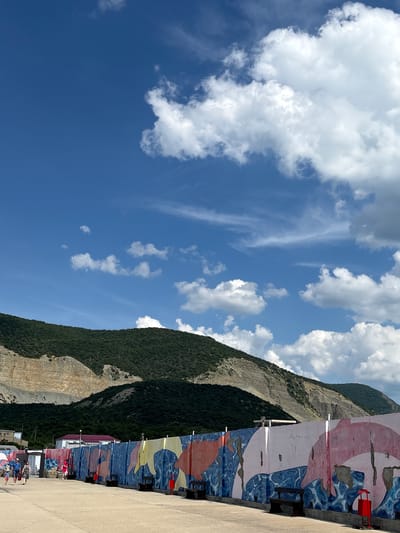 The image depicts an outdoor scene under a bright blue sky with scattered white clouds, characteristic of a sunny day. In the background, prominent, verdant mountains covered with dense green foliage rise steeply. A portion of one mountain face reveals exposed, lighter-colored rock or soil strata.

Stretching across the midground is a long, multi-sectioned concrete wall, serving as a barrier or fence. This wall is extensively decorated with vibrant murals, featuring large, abstract designs in shades of blue, red, yellow, and white/pink, which appear to represent stylized aquatic themes or marine life. Several dark-colored benches are placed at intervals along the base of this painted wall. Towards the right side of the wall, a distinctive red metal trash receptacle stands on the paved surface.

In the foreground, a wide, light-colored concrete or asphalt pavement extends horizontally, running parallel to the painted wall. On the left side of this pavement, several individuals are visible. A family group, consisting of a man, a woman, and a child, walks together. Further down the pavement to their right, another person, appearing to be a woman in a pink top, walks in the same direction. In the distance, behind the painted wall and nestled against the base of the mountains, a few modest buildings with reddish roofs can be discerned. The specific location is in the city of Bol'shoi Utrish, Russia.