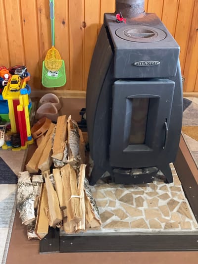 A top-down, slightly rotated indoor shot shows a wood-burning stove with a pile of birch logs, household objects, and toys. The stove is dark gray and positioned against a wall covered in mosaic tiles. To the left of the stove, against a wooden wall, there is a green and yellow fly swatter and a few white cups. At the bottom of the image, a pile of colorful plastic toys is visible. The overall setting suggests a domestic environment. The location in the city of Bibikha, Russia cannot be determined from the visual information provided.