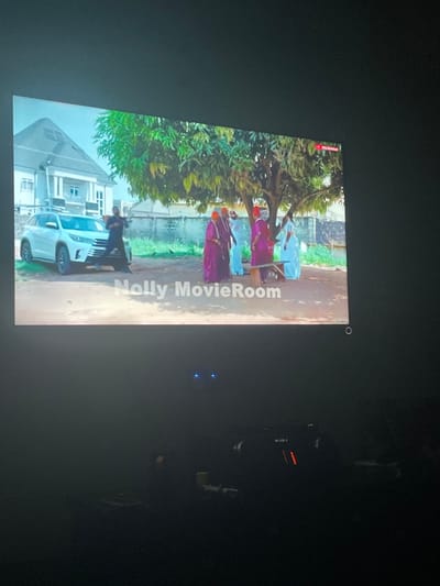 A scene from a film or show is displayed on a screen. Several people in traditional clothing stand under a tree next to a white vehicle and a building. Text is displayed vertically along the side of the screen. Other electronic devices are also visible. The room around the screen is dark.